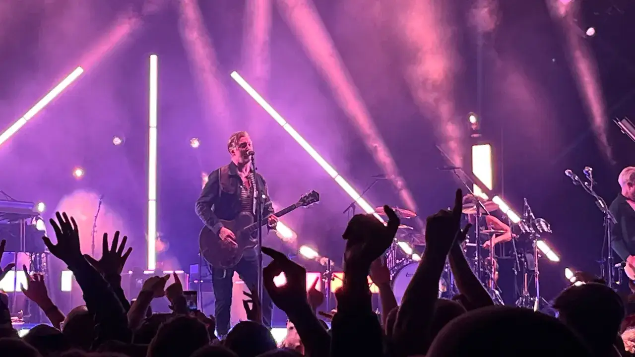 Concert des membres de Queens of the Stone Age sous des éclairages roses. La foule lève les mains en signe d'enthousiasme.