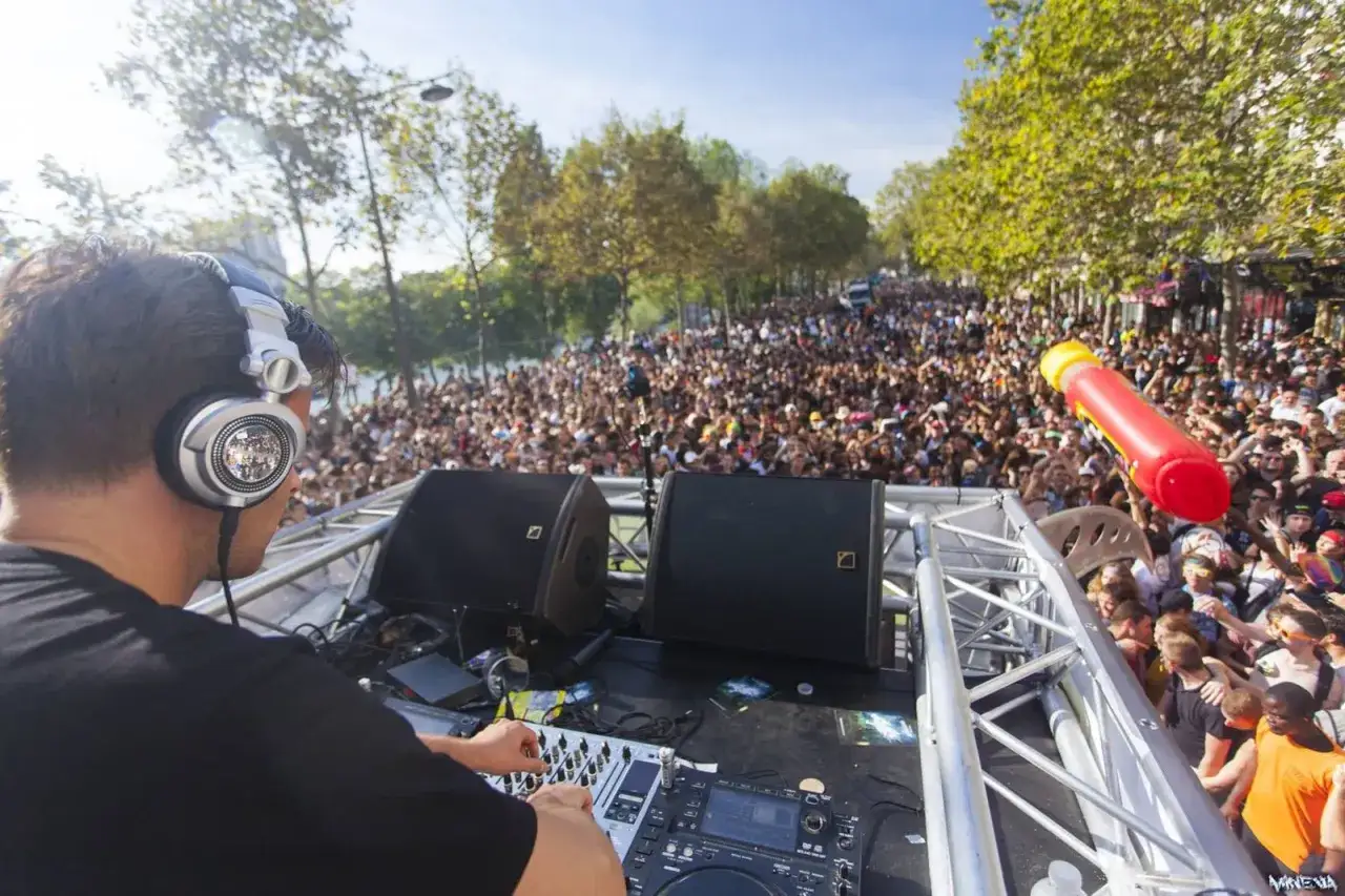 Un DJ &agrave; la mode mixe devant une foule immense, sous un ciel bleu. Une ambiance de f&ecirc;te en plein air.