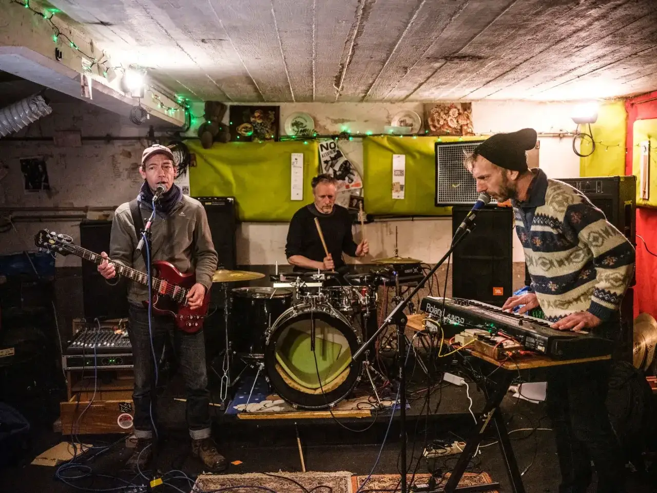 Trois musiciens jouent dans un local sombre, &eacute;clair&eacute; par des guirlandes vertes. Un guitariste chante, un batteur frappe sur sa batterie, et un clavi&eacute;riste joue. Ces oi boys sont en pleine action.