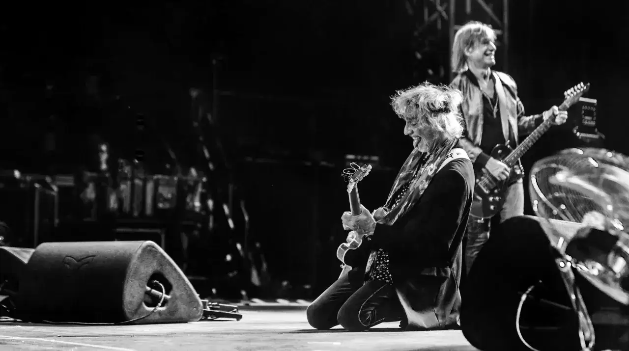 Un groupe français des années 70-80 en plein concert. Un guitariste s'agenouille, l'autre joue debout. L'énergie est palpable.