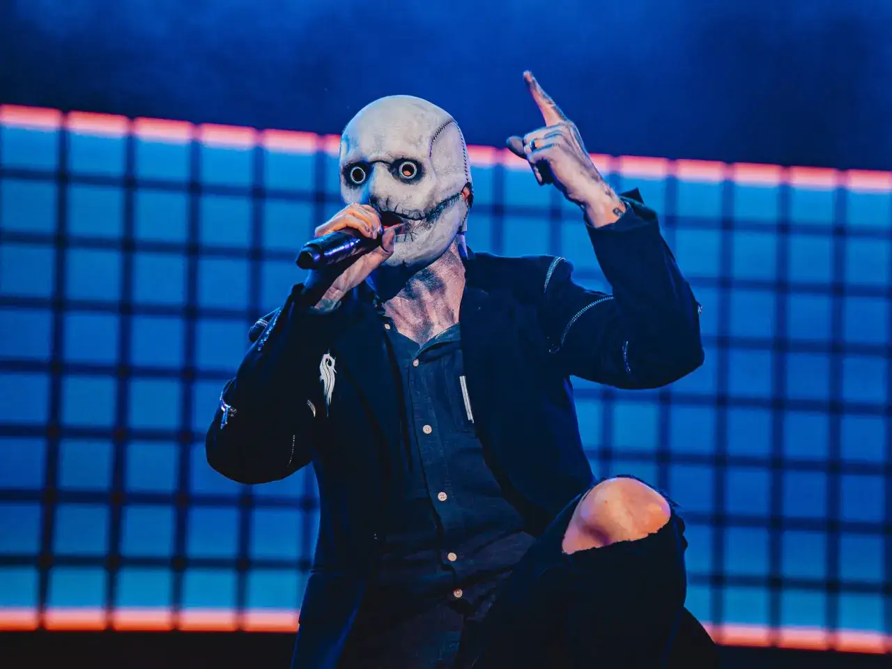 Le chanteur Slipknot, masqué et en pleine performance, pointe un doigt vers le ciel.