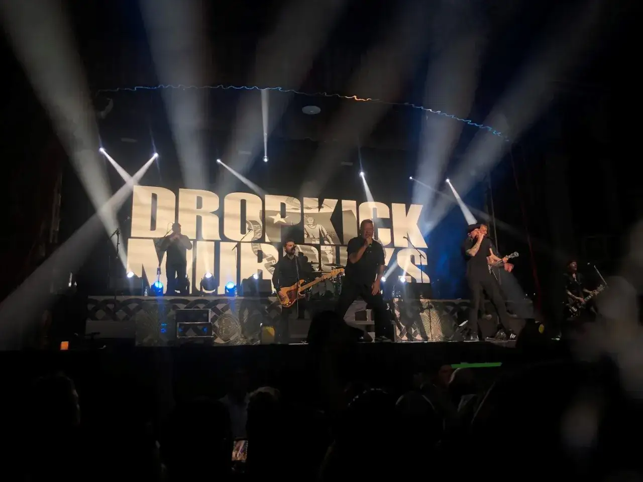 Les membres de Dropkick Murphys enflamment la scène sous les projecteurs. Le logo du groupe domine l'arrière-plan.