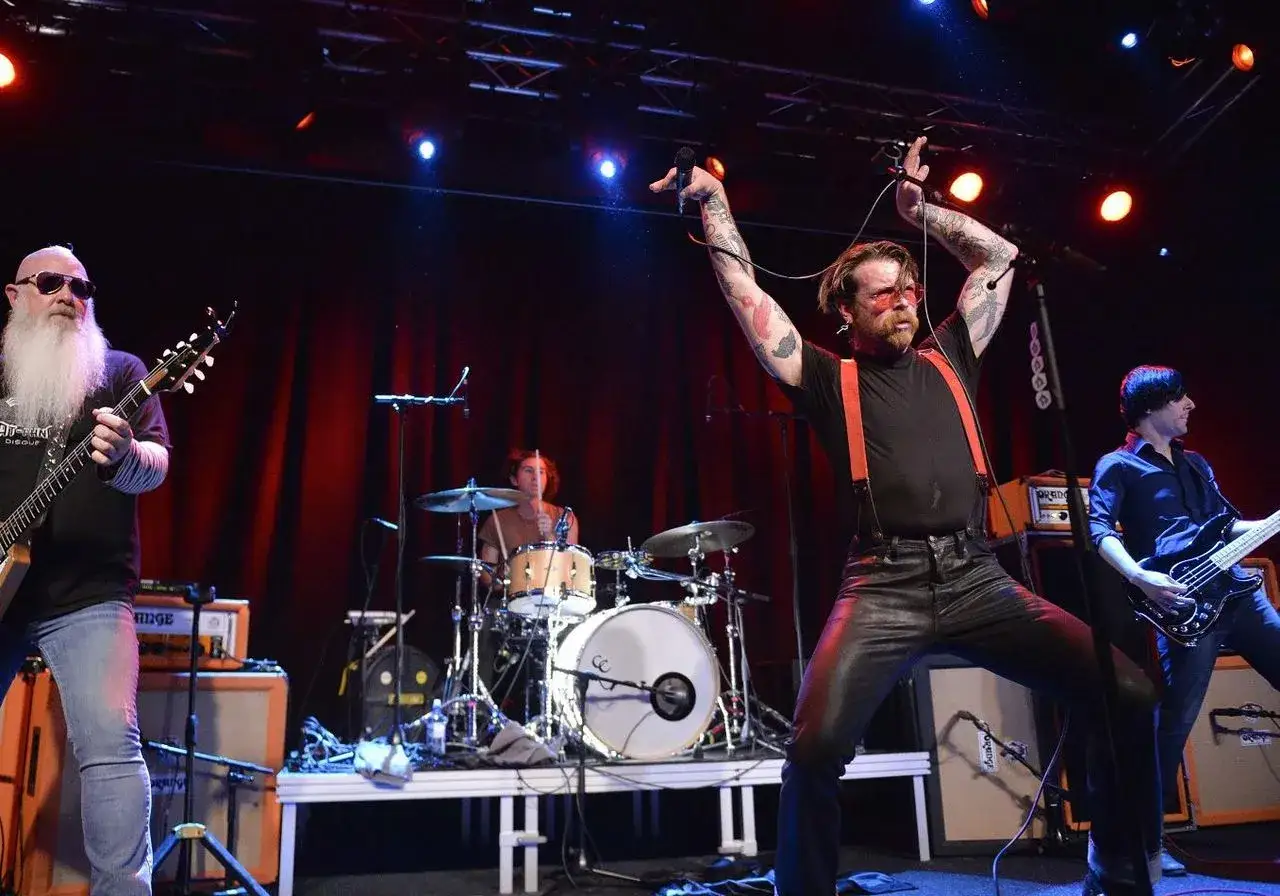 Les membres de Eagles of Death Metal en pleine performance. Le chanteur, torse nu, bras lev&eacute;s, est au centre. &Agrave; gauche, un guitariste &agrave; barbe blanche. &Agrave; droite, un bassiste. Au fond, un batteur.