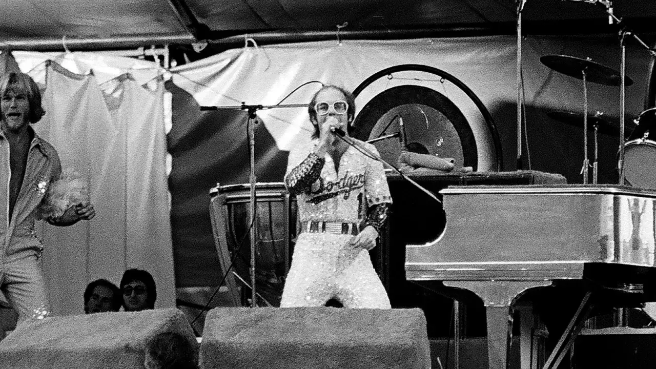 Elton John, un des 100 meilleurs groupes de rock de tous les temps, chante devant un piano et une batterie.