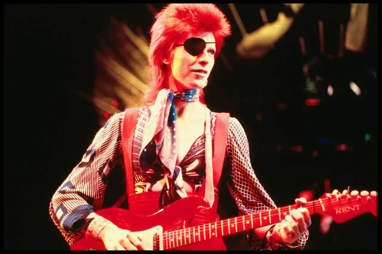 David Bowie, icône du rock, joue de la guitare rouge avec son groupe, arborant un bandeau sur l'œil et une chevelure flamboyante.