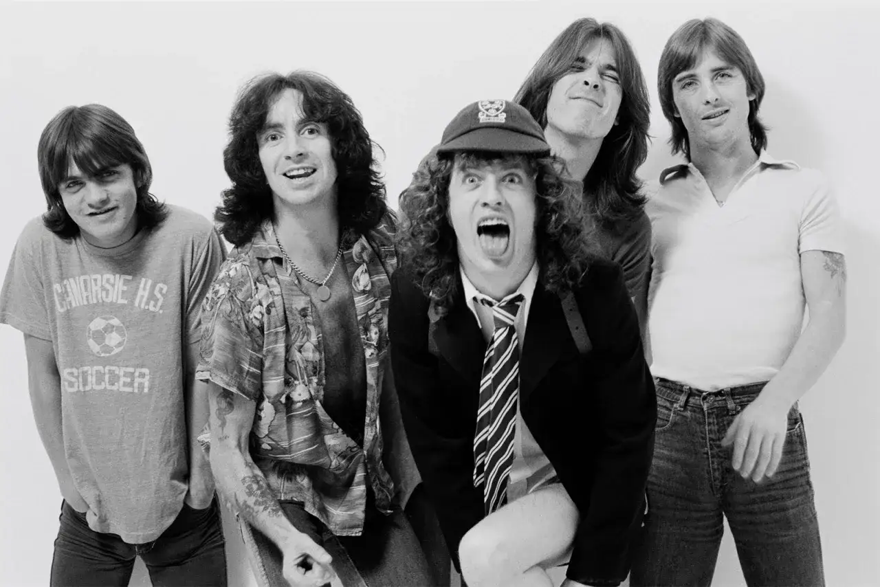 Le groupe ACDC pose, avec Bon Scott au centre, casquette et cravate, prêt à rugir. Les autres membres affichent des expressions variées.