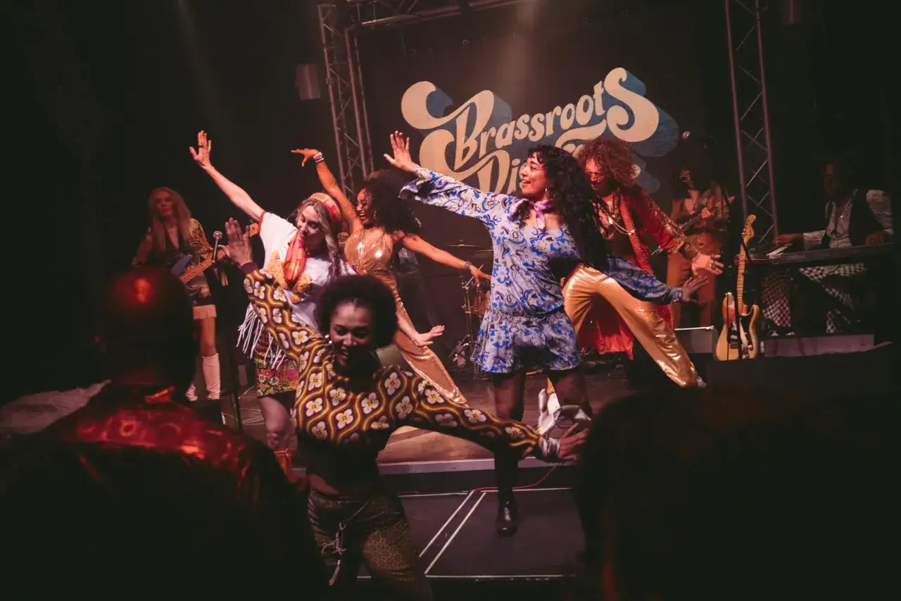 Un groupe de femmes, artistes funk, danse sur scène avec des tenues colorées et des pantalons brillants.
