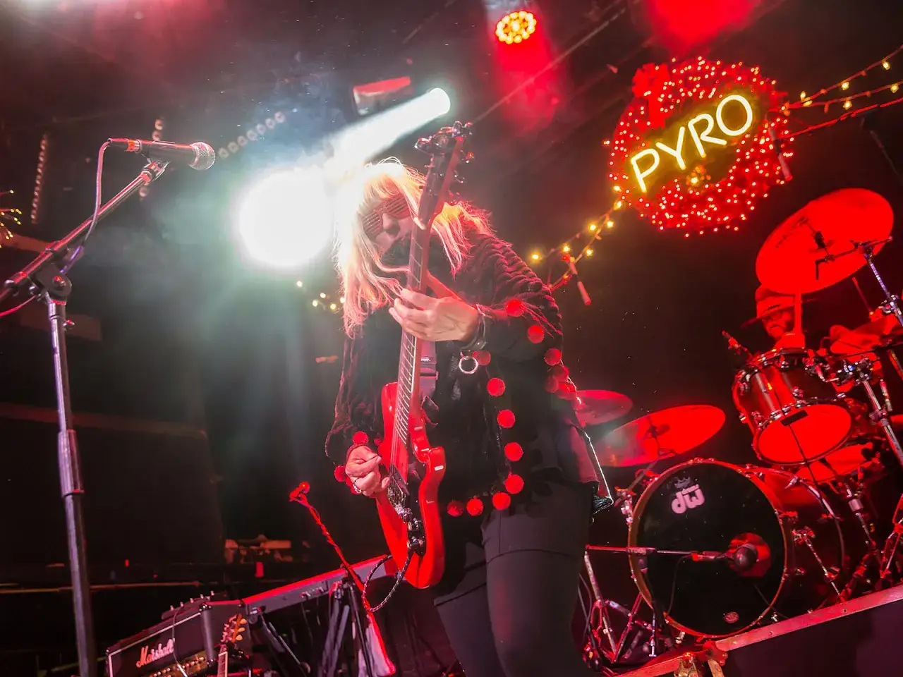 Une guitariste blonde, lunettes rouges, joue sur scène avec son groupe punk des années 90. Lumières vives, néon 