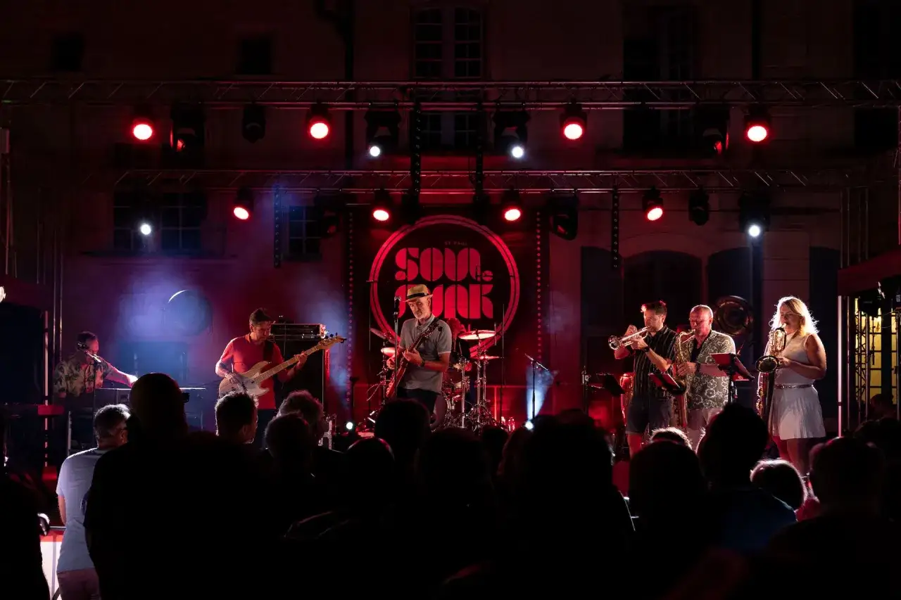 Un groupe de musique joue sur scène, éclairé par des projecteurs rouges. Un chanteur soul français, casquette sur la tête, joue de la guitare.