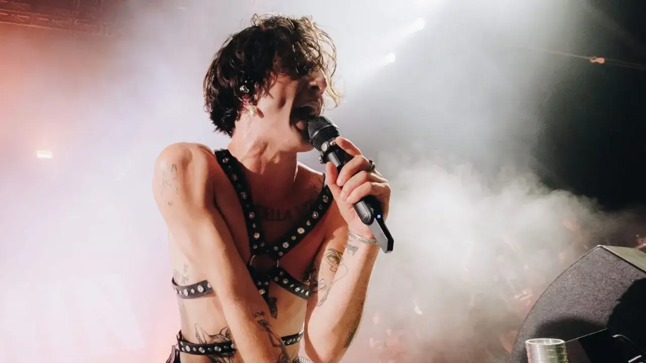 Le chanteur rock italien, torse nu et portant un harnais clouté, chante passionnément dans un microphone sur scène, entouré de fumée et de lumières.