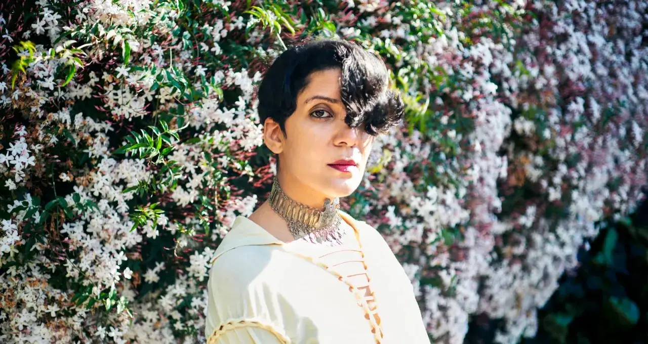 Fatima Al Qadiri, artiste à la chevelure courte, pose devant un mur de fleurs blanches et roses, parée d'un collier ethnique.