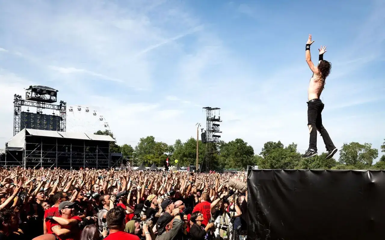 Un chanteur d'un groupe metal français connu saute au-dessus d'une foule en délire, sous un ciel bleu.