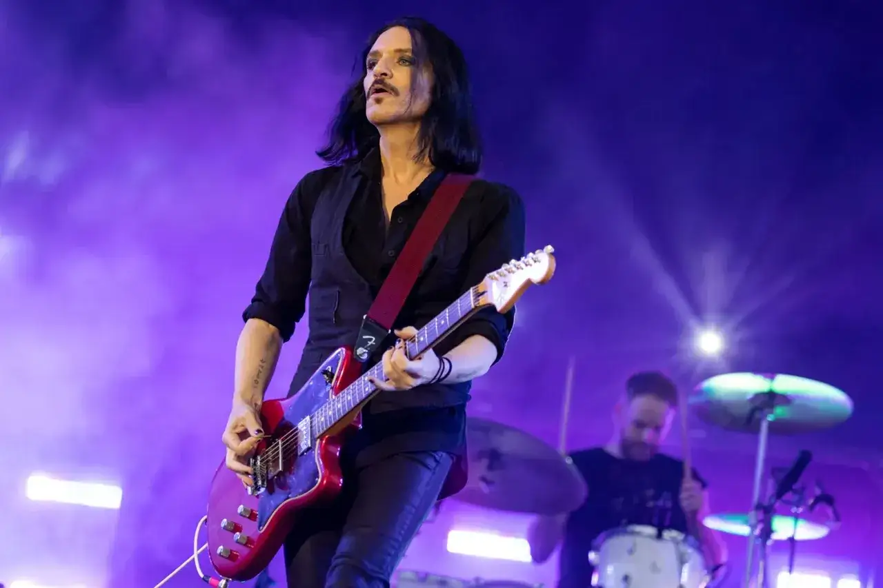 Brian Molko, le chanteur de Placebo, joue de la guitare rouge sur scène, sous une lumière violette.