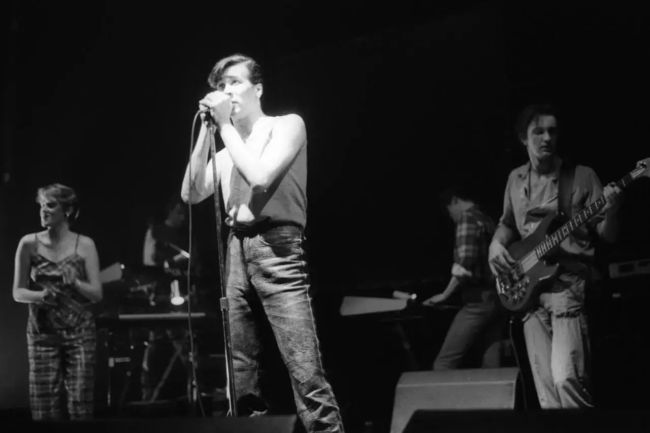 Un groupe rock anglais des ann&eacute;es 80 en pleine performance. Le chanteur, au premier plan, tient un micro. &Agrave; ses c&ocirc;t&eacute;s, un bassiste et d'autres musiciens sur sc&egrave;ne.