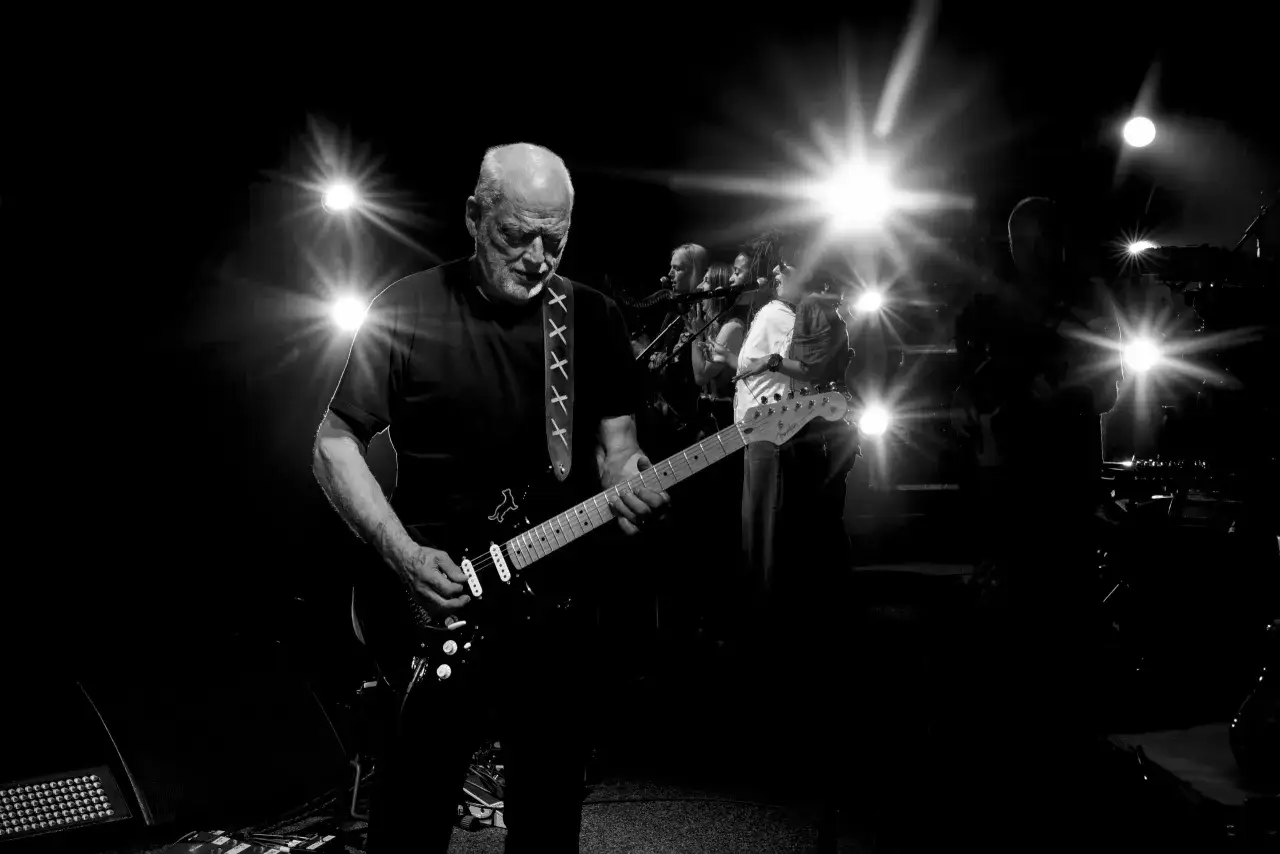 David Gilmour, icône du rock, joue de la guitare sur scène, entouré de musiciens. Une scène qui rappelle les grands albums de David Gilmour.