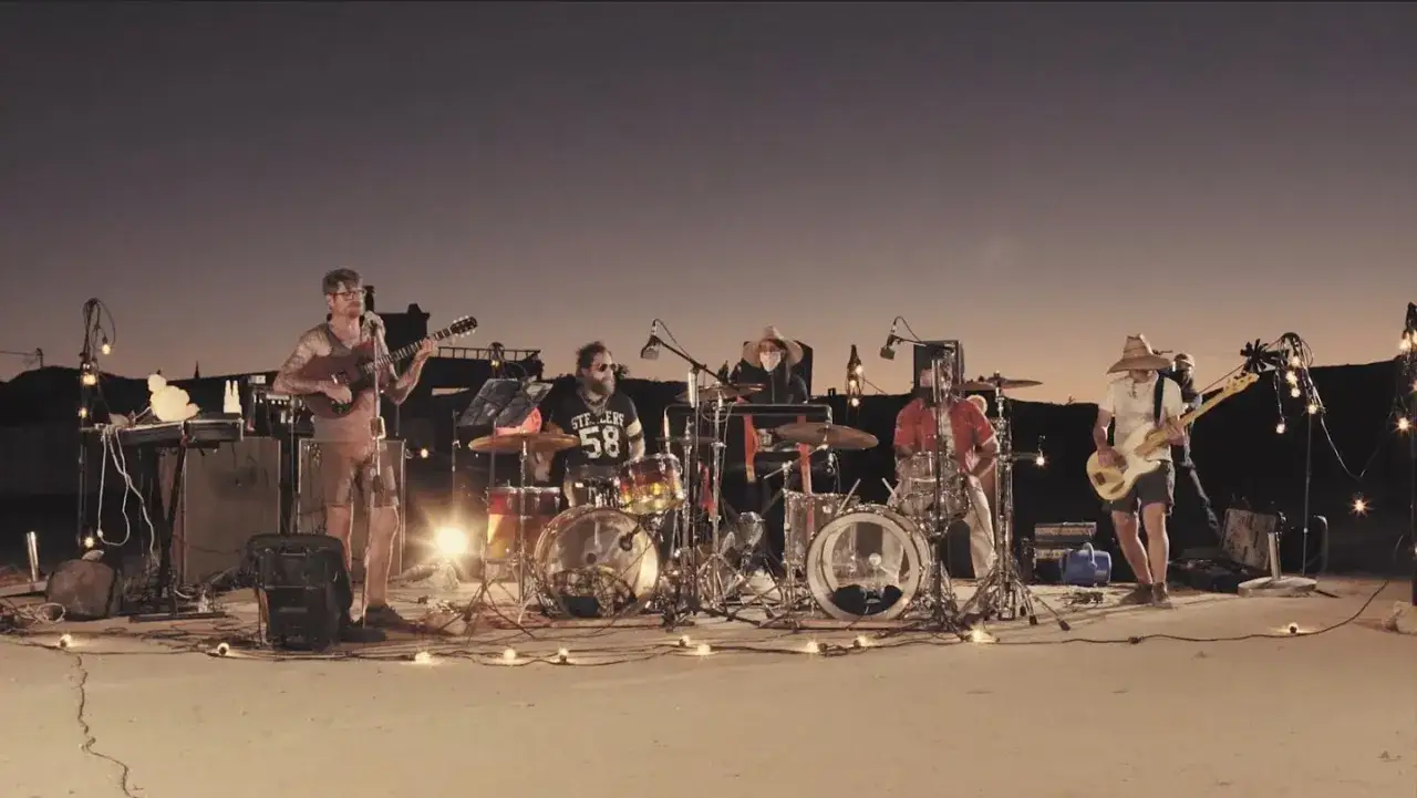Un groupe joue de la musique en plein air au coucher du soleil. Les membres du groupe, thee oh sees, sont entourés de lumières décoratives.