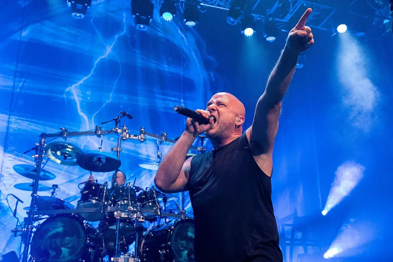 Le chanteur de Disturbed, David Draiman, hurle dans un micro, le bras lev&eacute;, sur sc&egrave;ne. Un batteur est visible derri&egrave;re lui.