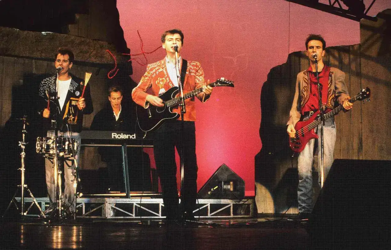 Un groupe australien des années 80 sur scène, avec des musiciens jouant de la guitare, de la basse et des claviers.