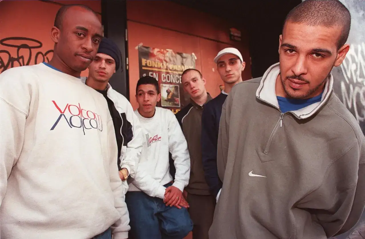 Un groupe de jeunes hommes, dont un ancien rappeur français, pose devant un mur orange.
