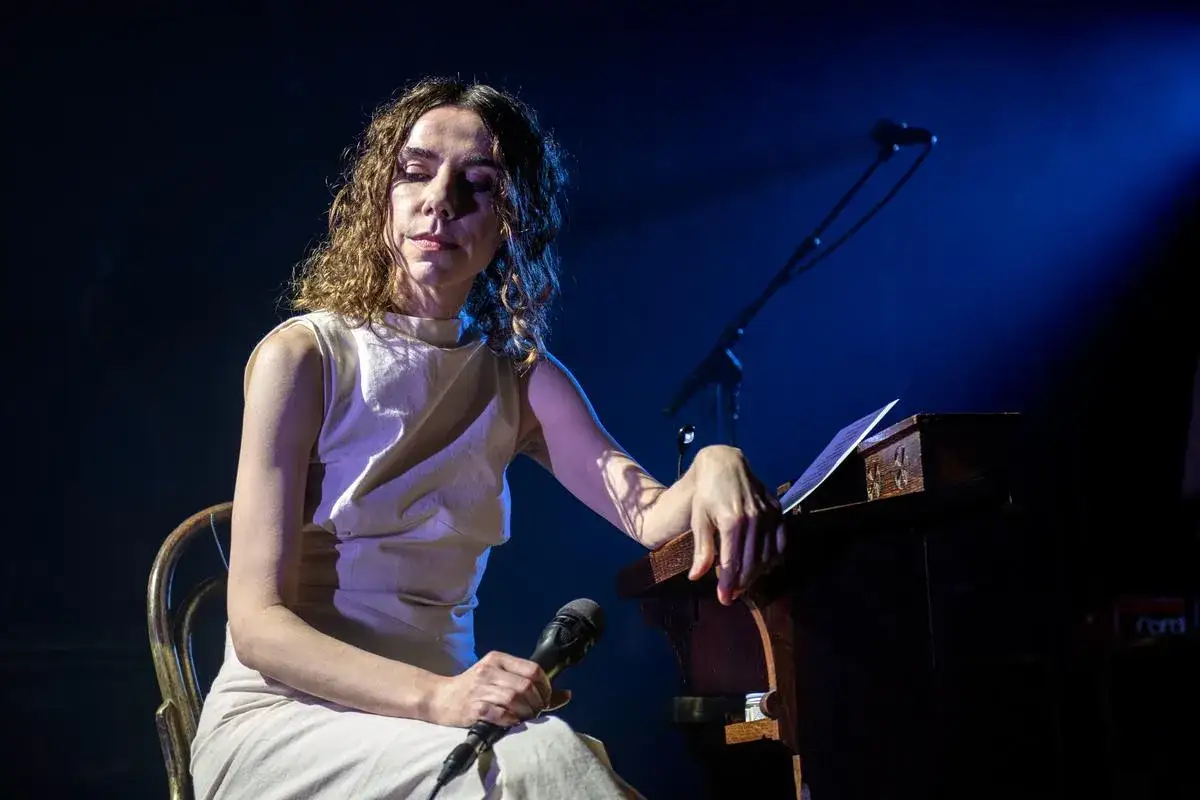 PJ Harvey, les yeux fermés, une main sur un pupitre en bois, l'autre tenant un micro, sur scène sous un éclairage bleuté.