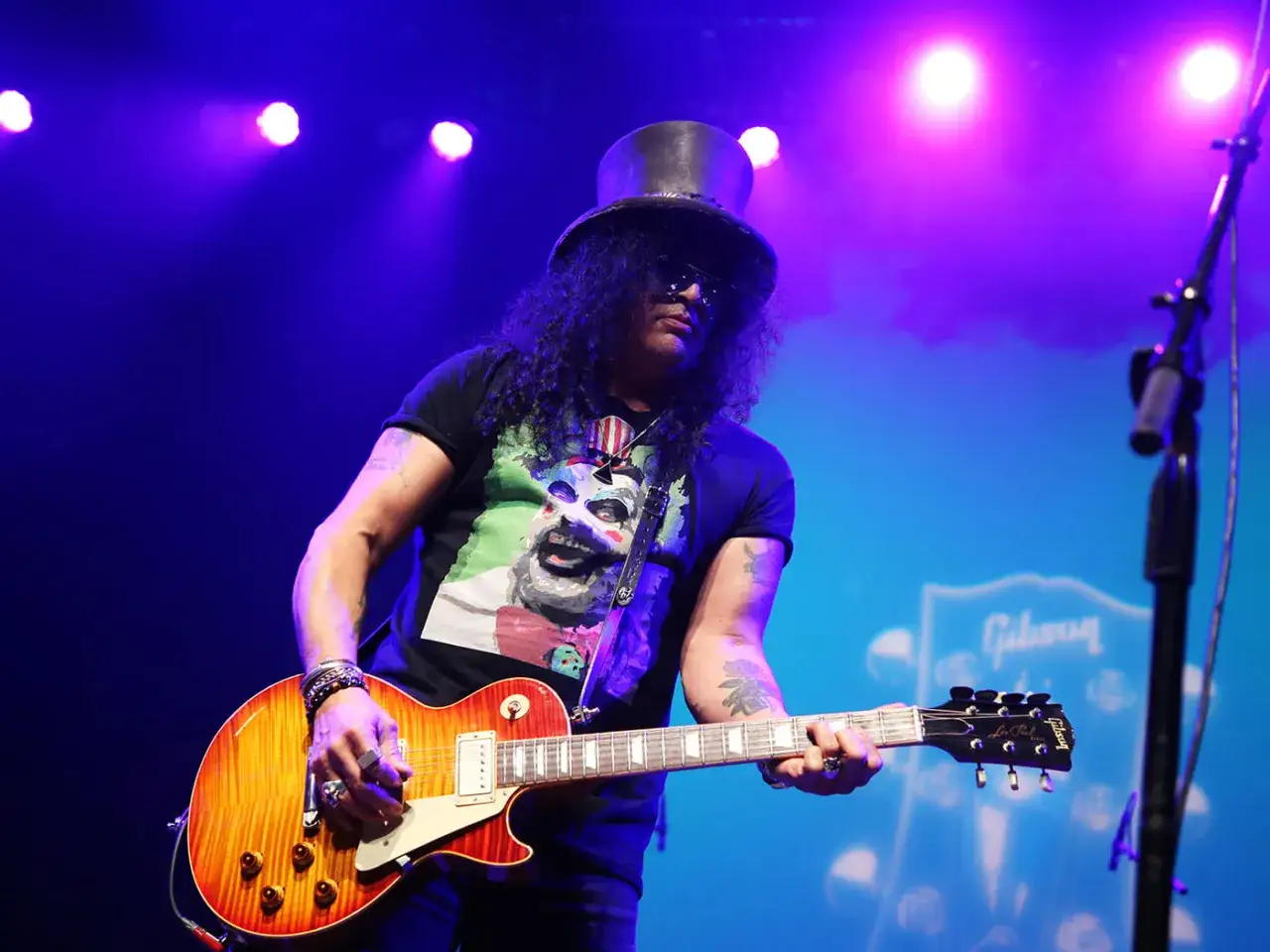 Le guitariste Slash, coiff&eacute; d'un chapeau haut-de-forme, joue sa Gibson sur sc&egrave;ne sous les lumi&egrave;res bleues.