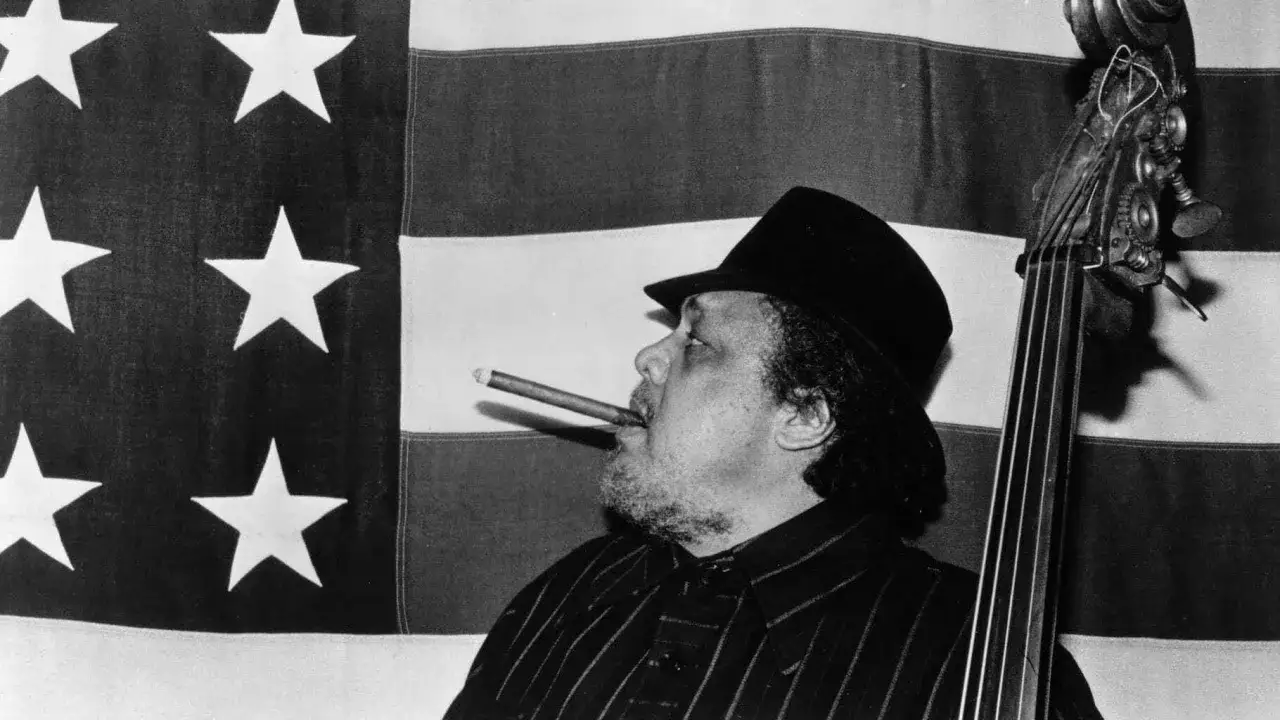 Charlie Mingus, le légendaire contrebassiste, fume un cigare devant le drapeau américain, prêt à jouer.