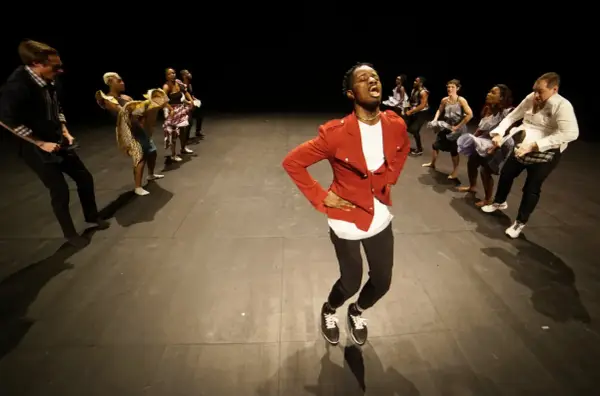 Un danseur afro 93, en veste rouge, mène une troupe sur scène. Les danseurs, aux tenues variées, exécutent des mouvements synchronisés.