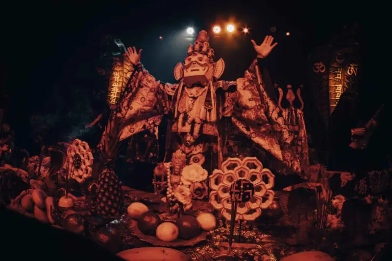 Un personnage masqué, drapé dans des tissus ornés, semble présider un rituel. Autour de lui, des offrandes de fruits, des serpents stylisés et des symboles sacrés évoquent un culte du feu mystérieux.