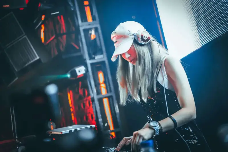 Une dj femme, casquette blanche et casque sur les oreilles, mixe dans une ambiance lumineuse et électrique.