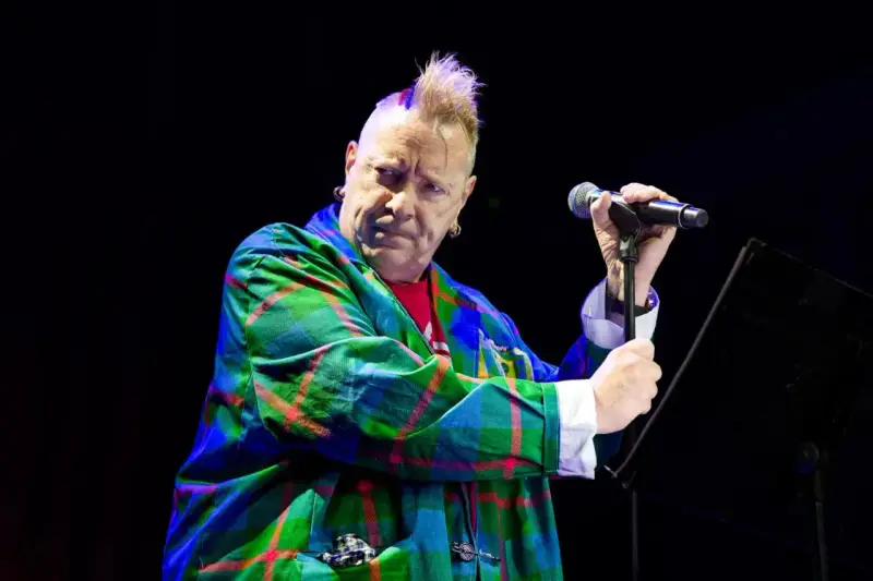 Le chanteur des Sex Pistols, John Lydon, avec sa crête iconique, porte une veste à carreaux écossais colorée et tient un microphone.