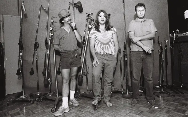 Trois hommes dans un studio d'enregistrement, entourés de micros. L'un d'eux, casquette et short, se touche la moustache. Husker du, un souvenir de cette époque.